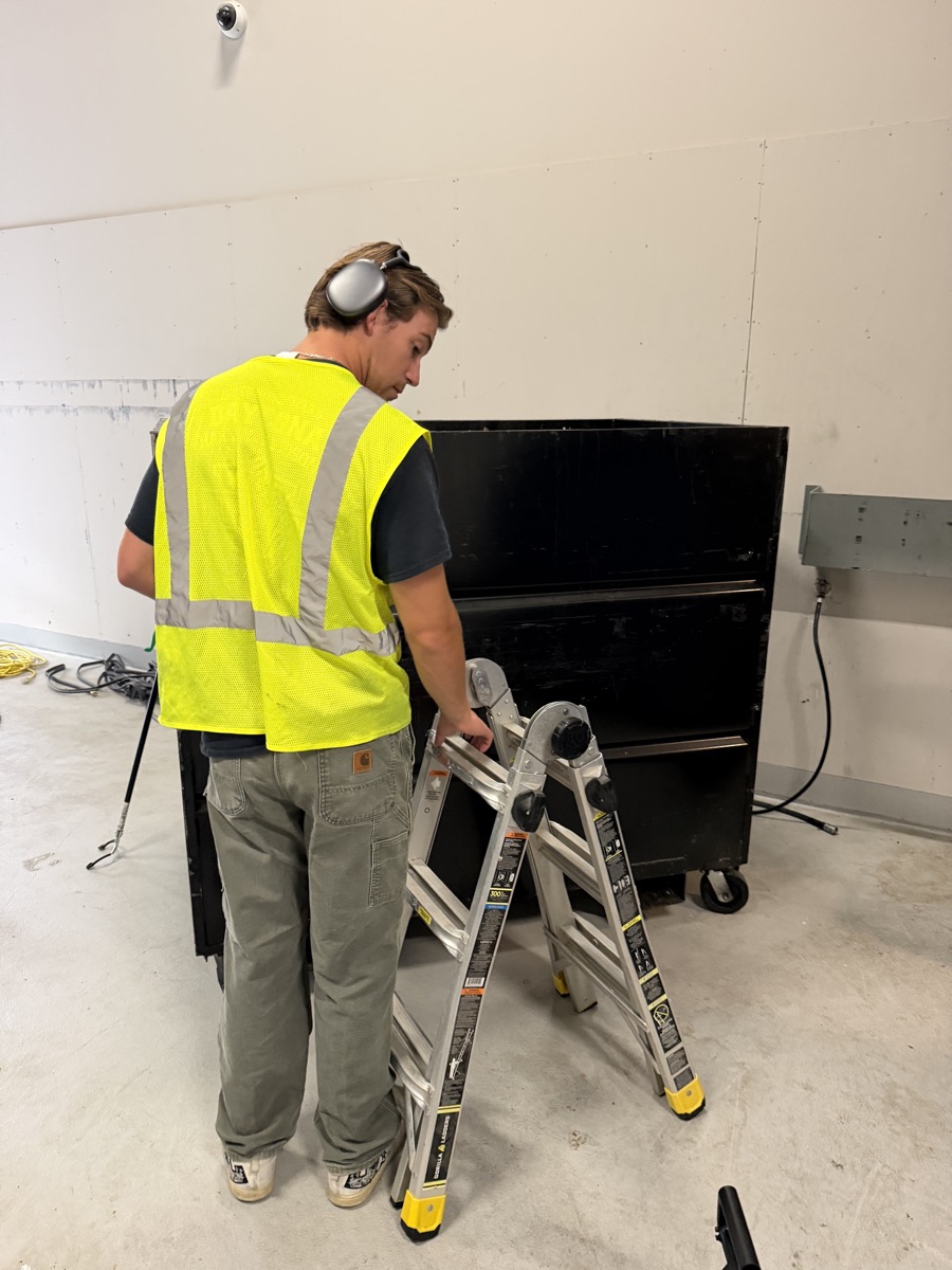 Logan, cleaning tech at Ventura Power Washing, in a safety vest setting up a ladder next to a dumpster
